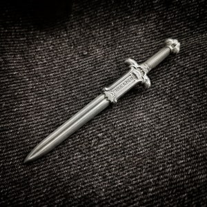 Könnte beinhalten: Ein silbernes Schwert mit filigranen Details und einem schwarzen Griff. Das Schwert liegt auf einem dunklen Jeansstoff.