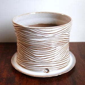 May include: A cylindrical ceramic planter with a white glaze and brown carved wavy line patterns. The planter features a matching circular saucer base with a small drainage hole visible at the bottom edge of the pot.