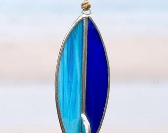 Op de afbeelding: Een surfboard ornament van glas-in-lood met een gespleten ontwerp van lichtblauw en donkerblauw glas, gevat in een zilverkleurig metalen frame met een kleine vin aan de onderkant. Het stuk hangt aan een touwtje van jute en bevat de tekst Tide Change.