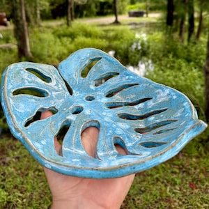 Könnte beinhalten: Eine handgefertigte Seifenschale aus Keramik in Form eines Monstera-Blattes. Die Schale ist hellblau mit weißer Marmorierung und braunen Rändern. Das Blatt hat mehrere Aussparungen. Der Hintergrund ist eine verschwommene Außenszene mit Bäumen und Grün.