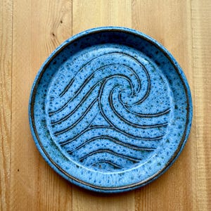 May include: A round, blue ceramic plate with a wave design. The plate has a speckled texture and a dark brown rim. The wave pattern is created with dark brown lines, adding contrast to the blue surface. The plate is on a wooden surface.