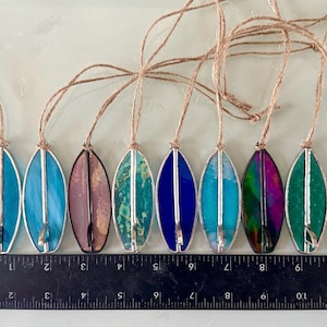 May include: A selection of stained glass surfboard decorations. Each ornament is about 10 cm long and features a different colour, including blue, purple, green, and pink, with a silver metal outline and a jute string for hanging.