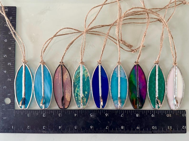 May include: A selection of stained glass surfboard decorations. Each ornament is about 10 cm long and features a different colour, including blue, purple, green, and pink, with a silver metal outline and a jute string for hanging.