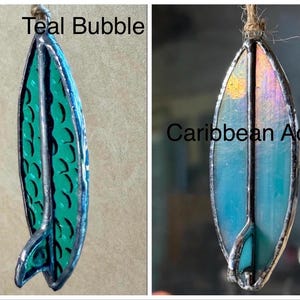 May include: Two stained glass surfboard ornaments. One is teal with a bubble pattern and the text "Teal Bubble". The other is Caribbean blue with a sun design and the text "Caribbean Aqua". Both have a silver border and a twine loop.