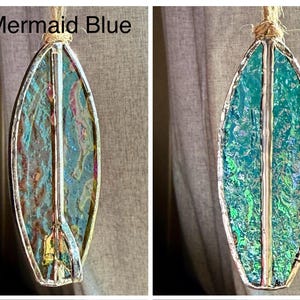 May include: A stained glass surfboard ornament in shades of teal, blue, and gold, with a silver-coloured metal border. The ornament is suspended by a natural fibre cord. The text "Mermaid Blue" is visible in the image.