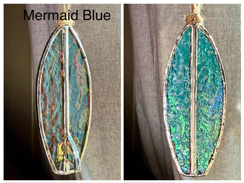 May include: A stained glass surfboard ornament in shades of teal, blue, and gold, with a silver-coloured metal border. The ornament is suspended by a natural fibre cord. The text "Mermaid Blue" is visible in the image.