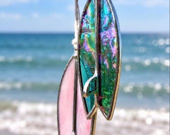 May include: Two surfboard-shaped ornaments made of stained glass with metal soldering. One surfboard is solid light pink, while the other features a vibrant, iridescent green and purple finish. Both pieces hang from white string loops.