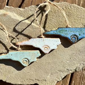 May include: Three ceramic ornaments shaped like the state of North Carolina, each with a unique glaze in shades of blue, white, and green. They feature a spiral design and are suspended by twine. The ornaments are displayed on a textured stone surface.