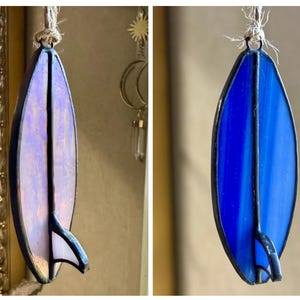 May include: Stained glass surfboard ornament in shades of blue and purple, with a black outline and fin. The ornament is suspended by a natural fibre cord. A decorative piece for a beach or surf theme.