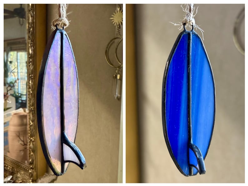 May include: Stained glass surfboard ornament in shades of blue and purple, with a black outline and fin. The ornament is suspended by a natural fibre cord. A decorative piece for a beach or surf theme.