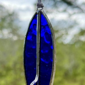 May include: A surfboard-shaped stained glass suncatcher in a deep blue colour. The glass is framed in silver-coloured metal, with a vertical metal accent. A white cord is attached for hanging.