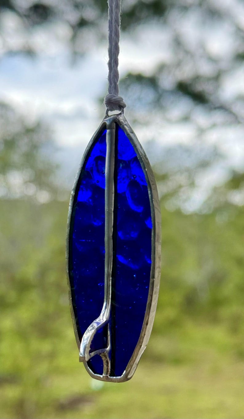May include: A surfboard-shaped stained glass suncatcher in a deep blue colour. The glass is framed in silver-coloured metal, with a vertical metal accent. A white cord is attached for hanging.