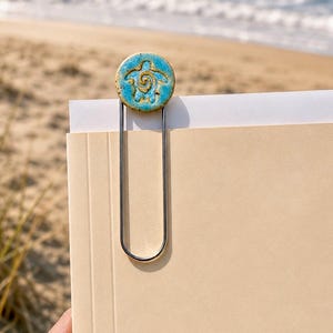 May include: A large metal paperclip featuring a circular, textured ceramic topper in a vibrant turquoise blue glaze. The topper displays a debossed, golden-brown sea turtle motif at its center. The paperclip is attached to a stack of beige paper.