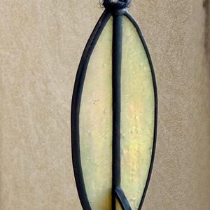 May include: An oval-shaped stained glass pendant with a black outline and a central black stripe. The glass has a yellow and gold iridescent finish. The text "Golden Tide Black" is at the bottom of the image.