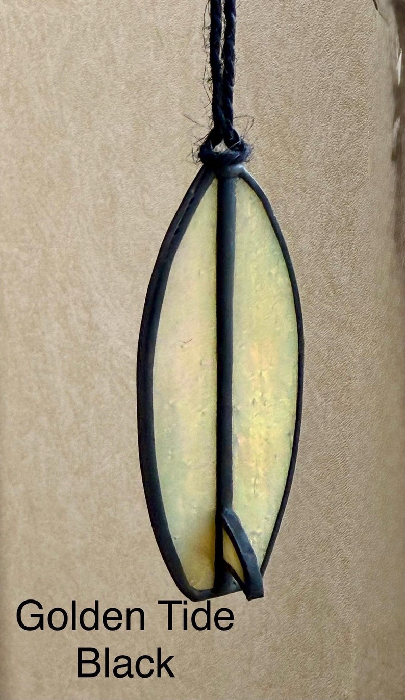 May include: An oval-shaped stained glass pendant with a black outline and a central black stripe. The glass has a yellow and gold iridescent finish. The text "Golden Tide Black" is at the bottom of the image.