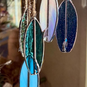 May include: A collection of five stained glass surfboard ornaments hanging from jute twine. Each surfboard features a unique color, including iridescent white, deep navy blue with sparkles, teal, light blue, and green, all framed in silver metal.