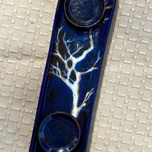 Puede incluir: Una bandeja rectangular de cerámica azul oscuro con dos hendiduras circulares y un diseño de árbol central. El árbol es blanco con detalles marrones. La bandeja mide aproximadamente 30 cm de largo.
