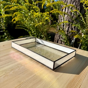 Może przedstawiać: Prostokątna taca ze szkła witrażowego z białą obwódką i czarnym wykończeniem. Taca ma przezroczystą, teksturowaną szklaną podstawę. Taca stoi na jasnej drewnianej powierzchni z naturalnym tłem.