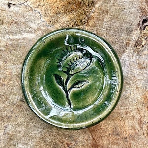 May include: A round, glossy ceramic dish in a deep forest green glaze. The center features a debossed, dark-outlined illustration of a Venus flytrap plant with its jaw-like leaves open, showcasing sharp, tooth-like edges and a central stem.