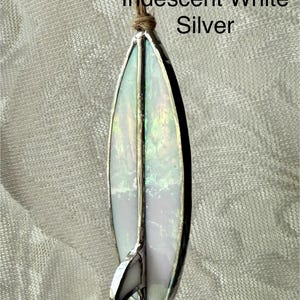May include: Iridescent white and silver stained glass surfboard ornament. The surfboard has a silver border and a small fin. The text "Iridescent White Silver" is above the surfboard. A brown string is attached to the top.