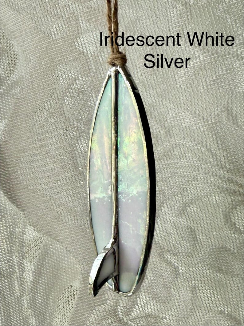 May include: Iridescent white and silver stained glass surfboard ornament. The surfboard has a silver border and a small fin. The text "Iridescent White Silver" is above the surfboard. A brown string is attached to the top.