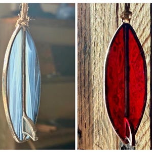 May include: Two stained glass surfboard ornaments. One is blue and white, the other is red. Both have a metal frame and are suspended by a natural fibre cord. The ornaments are designed to resemble surfboards.