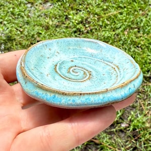 Puede incluir: Un pequeño plato redondo de cerámica con un esmalte turquesa y un diseño en espiral en el centro. El borde tiene un borde marrón. El plato se sostiene en una mano sobre un fondo de hierba verde.