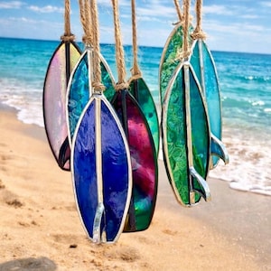 May include: A selection of colourful stained glass surfboard ornaments suspended by twine. The surfboards display shades of blue, green, pink, and purple, with silver metal edging and fins. The background shows a sandy beach and the ocean.