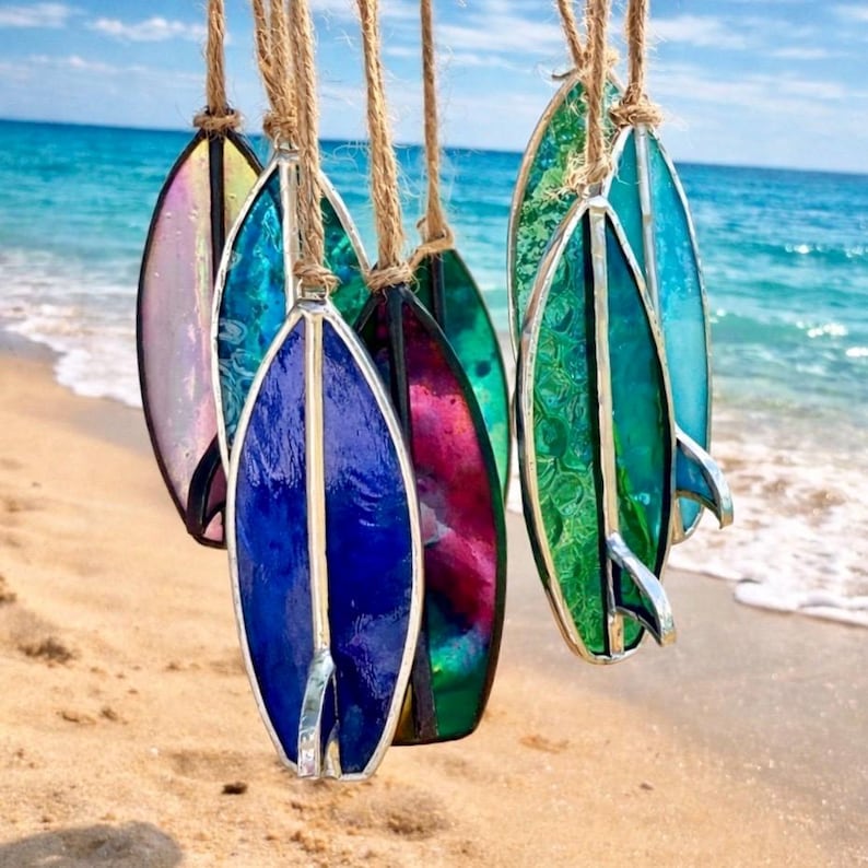 May include: A selection of colourful stained glass surfboard ornaments suspended by twine. The surfboards display shades of blue, green, pink, and purple, with silver metal edging and fins. The background shows a sandy beach and the ocean.