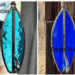 May include: Two stained glass surfboard ornaments. One is turquoise with the text "Blue Bubble", the other is deep blue with the text "Deep Sea". Both have a silver metal border and a twine loop for hanging.