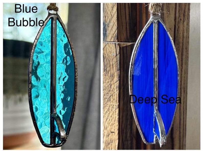 May include: Two stained glass surfboard ornaments. One is turquoise with the text "Blue Bubble", the other is deep blue with the text "Deep Sea". Both have a silver metal border and a twine loop for hanging.