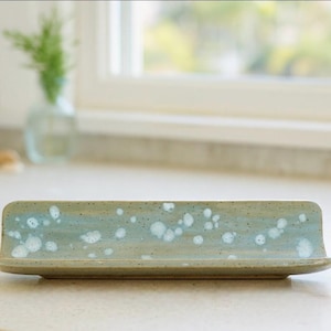 Peut inclure: Un beurrier rectangulaire en céramique avec un glaçage tacheté bleu clair et vert. Le plat a une forme légèrement incurvée et un petit rebord sur le devant. L'arrière-plan comprend une fenêtre et un vase avec de la verdure.