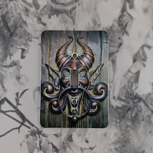 May include: A decorative light switch cover with a horned demon face, large ears, and a ring-shaped handle. The design uses brown, bronze, and green tones, set against a wood-grain background. The switch opening is a vertical rectangle.