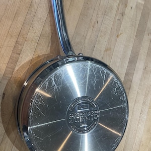 May include: A stainless steel Farberware Millennium skillet with a long, polished handle. The underside of the pan features a circular stamped logo that reads Farberware Millennium, along with text indicating stainless steel construction and design origin.