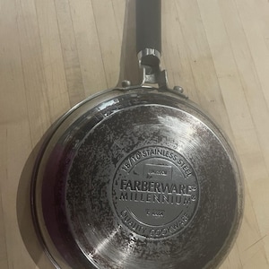 May include: Stainless steel Farberware Millennium skillet with a black handle. The bottom of the pan is marked "18/10 Stainless Steel" and "Quality Cookware". The pan is approximately 8 inches in diameter.