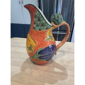 May include: A ceramic pitcher with a vibrant orange background featuring painted illustrations of yellow peppers, purple eggplants, and green artichoke leaves. The handle is topped with a sculpted green artichoke finial.