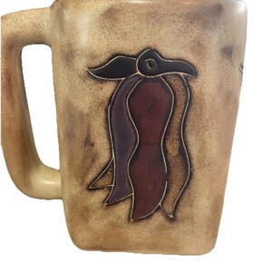 May include: A beige ceramic mug with a rectangular shape and a large handle. The mug features a decorative design of a stylized flower in shades of brown, purple, and black. The mug has a textured surface.