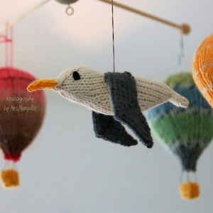 Könnte beinhalten: Ein gestricktes Möwen-Mobile mit weißem Körper, grauen Flügeln und einem gelben Schnabel. Die Möwe hängt an einem Holzarm eines Mobiles mit vier Heißluftballons in verschiedenen Farben und Mustern.