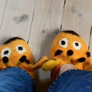 Op de afbeelding: Een paar oranje en bruine gebreide pantoffels in de vorm van een giraffe met zwarte ogen en een bruine neus. De pantoffels worden gedragen door iemand die een blauwe spijkerbroek draagt. De tekst "Knitography by Mrs. Humpitz" is zichtbaar in de rechterbenedenhoek van de afbeelding.