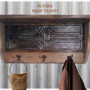 May include: Rustic wooden wall shelf with a tin back panel and three hooks. The shelf has a distressed brown finish and the tin panel has a silver and black pattern. The shelf is mounted on a white corrugated metal wall.