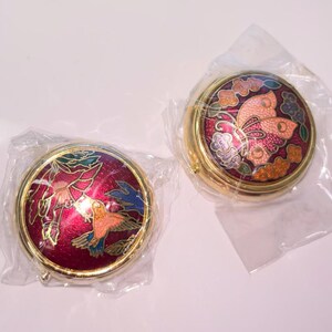 Op de afbeelding: Twee ronde, goudkleurige, decoratieve dozen met scharnierende deksels. De deksels hebben kleurrijke emaille-ontwerpen. E&eacute;n doos heeft een vlinderontwerp met roze, oranje en paarse accenten. De andere doos heeft een vogelontwerp met rode, oranje en blauwe accenten.