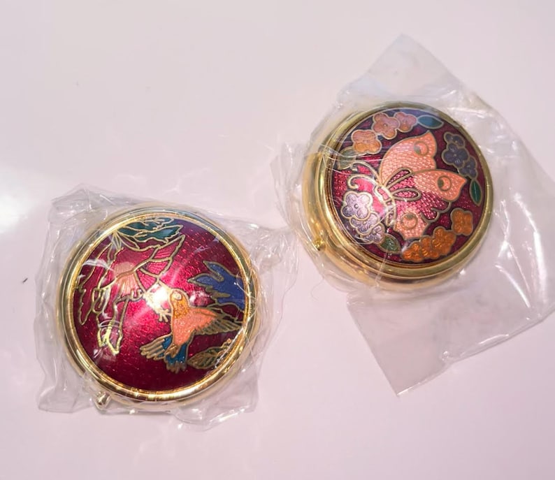 Op de afbeelding: Twee ronde, goudkleurige, decoratieve dozen met scharnierende deksels. De deksels hebben kleurrijke emaille-ontwerpen. E&eacute;n doos heeft een vlinderontwerp met roze, oranje en paarse accenten. De andere doos heeft een vogelontwerp met rode, oranje en blauwe accenten.