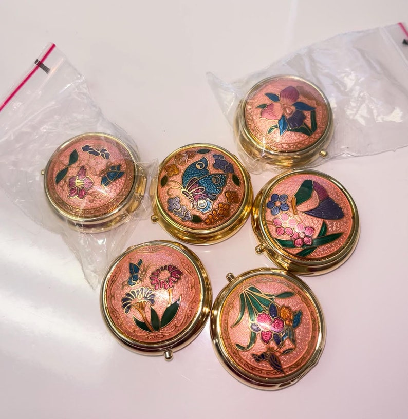 Op de afbeelding: Zes kleine ronde gouden sieradendoosjes met roze deksels en kleurrijke bloemen- en vlindermotieven.