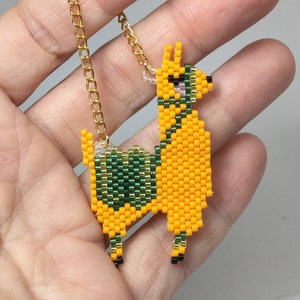 May include: A yellow llama charm with green and pink details, hanging from a gold chain. The charm is made of beads and is a cute and whimsical accessory.