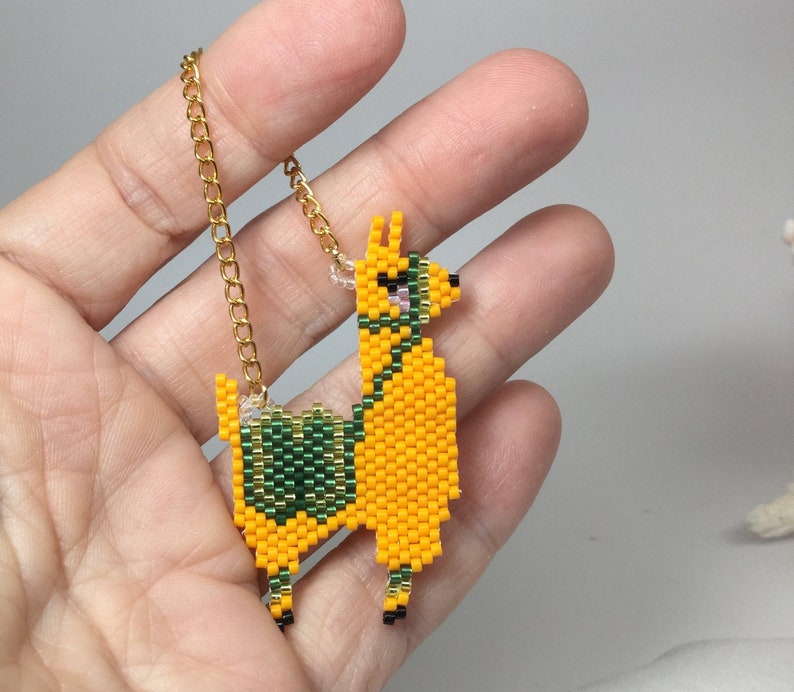 May include: A yellow llama charm with green and pink details, hanging from a gold chain. The charm is made of beads and is a cute and whimsical accessory.