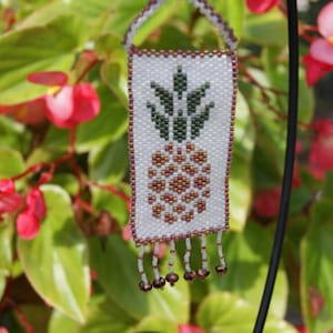 May include: A beaded pineapple keychain with a white background, green leaves, and brown pineapple details. The keychain is hanging from a black metal hook.