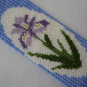 May include: A beaded bracelet with a white background and a purple and green flower design.