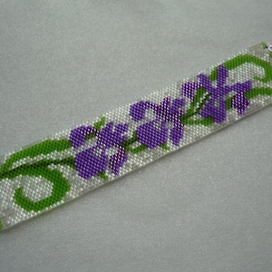 Może przedstawiać: Biała bransoletka z koralików z fioletowym i zielonym wzorem kwiatowym. Bransoletka jest wykonana z koralików nasiennych i ma srebrne zapięcia.