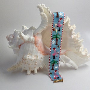 Può includere: Un braccialetto di perline blu e rosso con un design natalizio con fenicotteri rosa, palme, fiocchi di neve e cappelli di Babbo Natale. Il braccialetto è appoggiato su una conchiglia bianca.