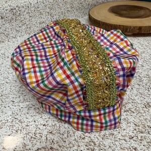 May include: A colorful turban with a plaid pattern in red, yellow, green, and blue. The turban is adorned with a gold-colored, ornate trim. The fabric is gathered and folded, creating a textured appearance.